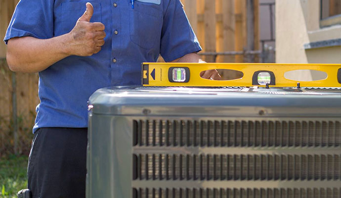 HVAC technician in a blue uniform giving a thumbs up while using a level tool on an outdoor air conditioning unit