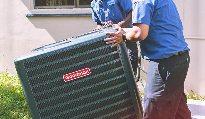 Two HVAC technicians in blue uniforms working together to move and install a Goodman air conditioning unit outdoors