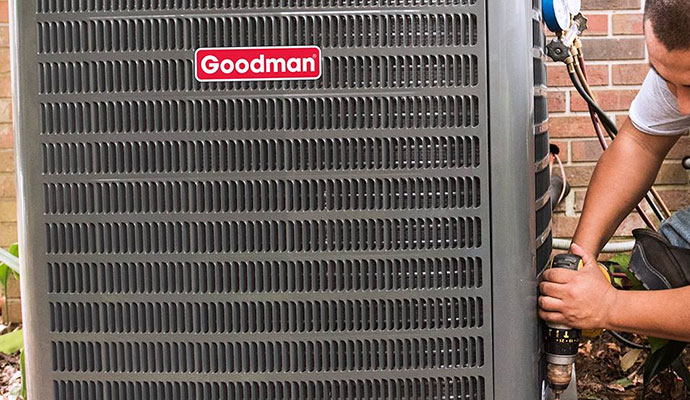 A professional technician using a power drill to service a Goodman air conditioning unit outside a brick home
